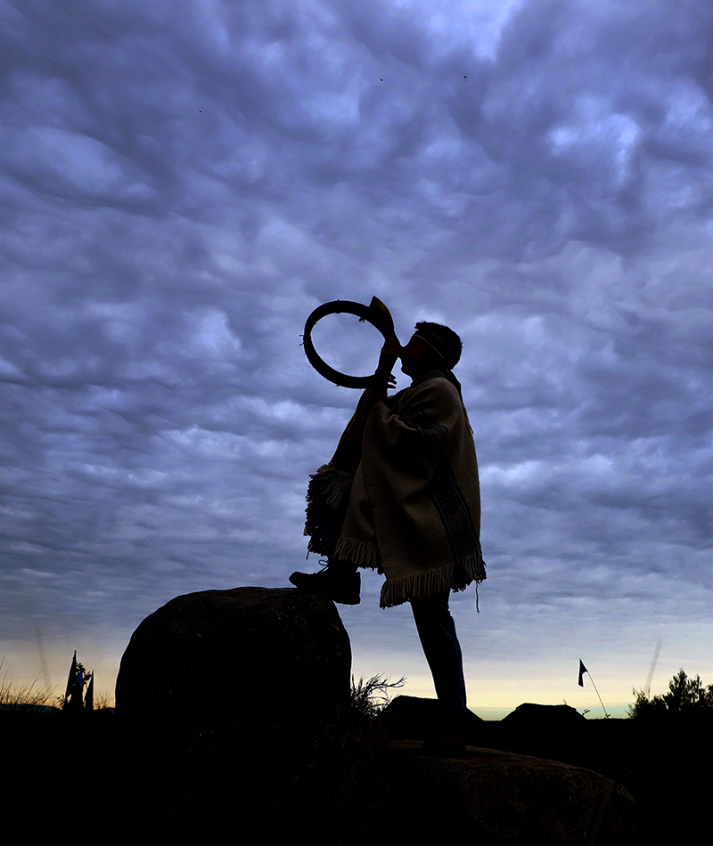 Silueta de un músico mapuche soplando un instrumento ancestral en lo alto de una roca en Peñalolén, bajo un cielo nublado al atardecer